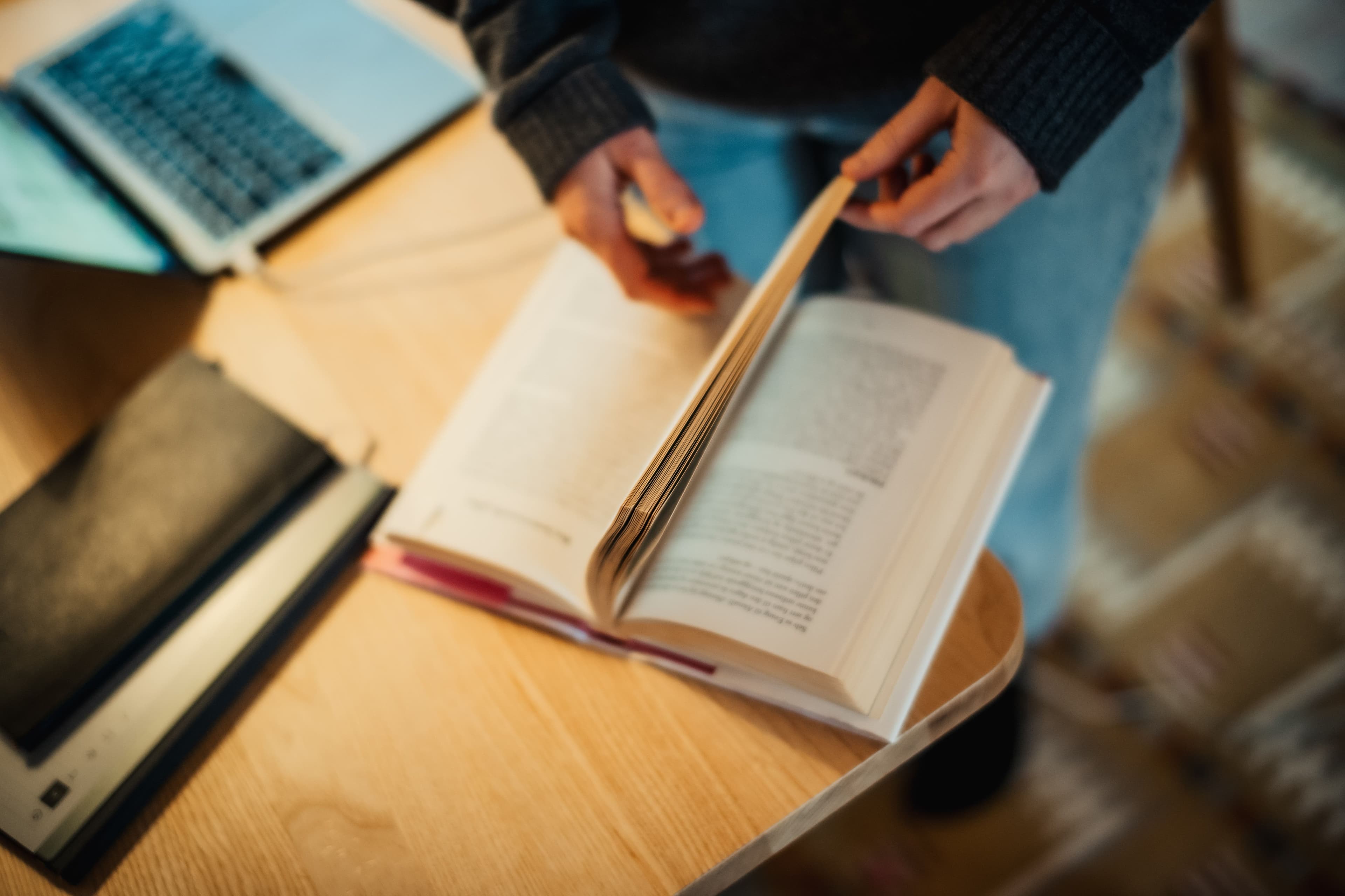 Skrivebok med bok, PC og notatblokk. En kvinne blar i boka.